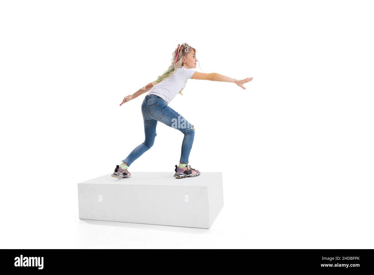 Full-length portrait of smiling girl in casual clothes standing on big box isolated on white studio background. Happy childhood concept. Stock Photo