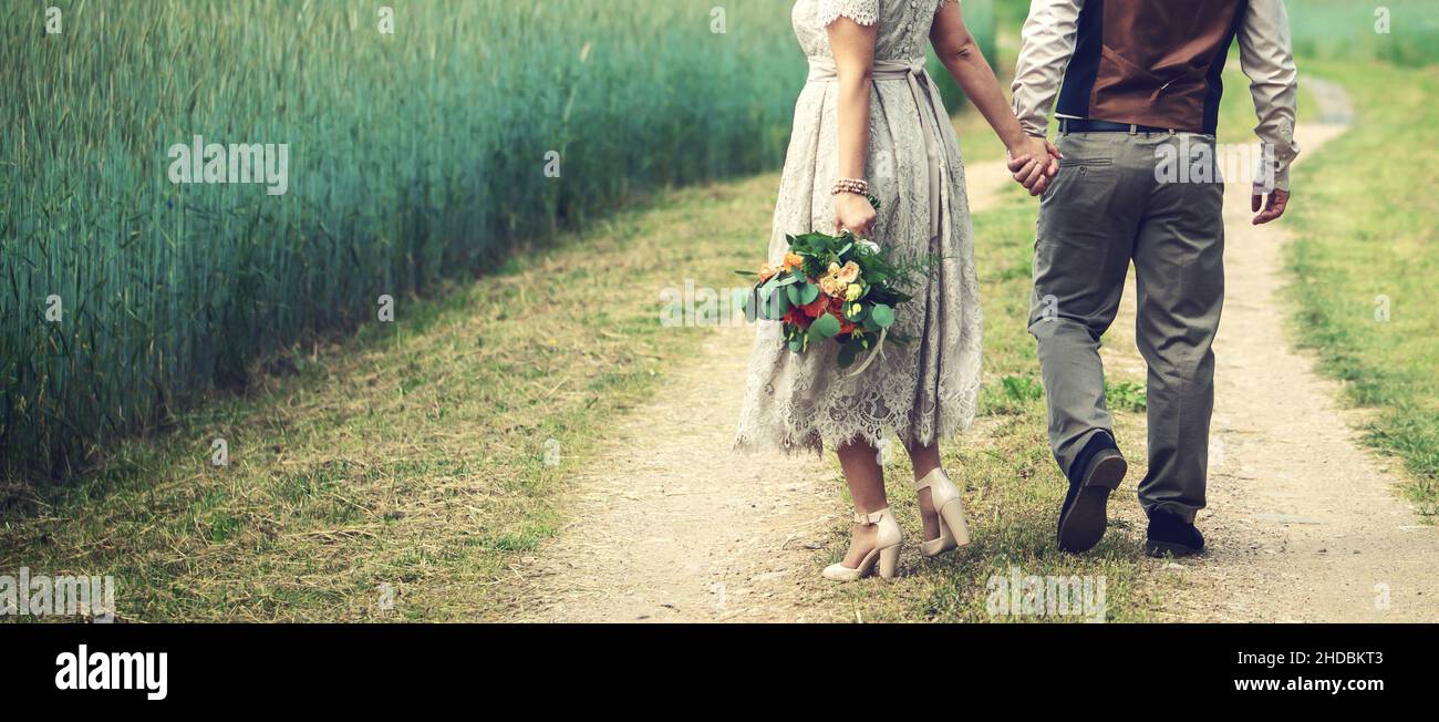 wedding couple, bride and groom holding hands and walks the country road beside cereal field. back view copy space Stock Photo