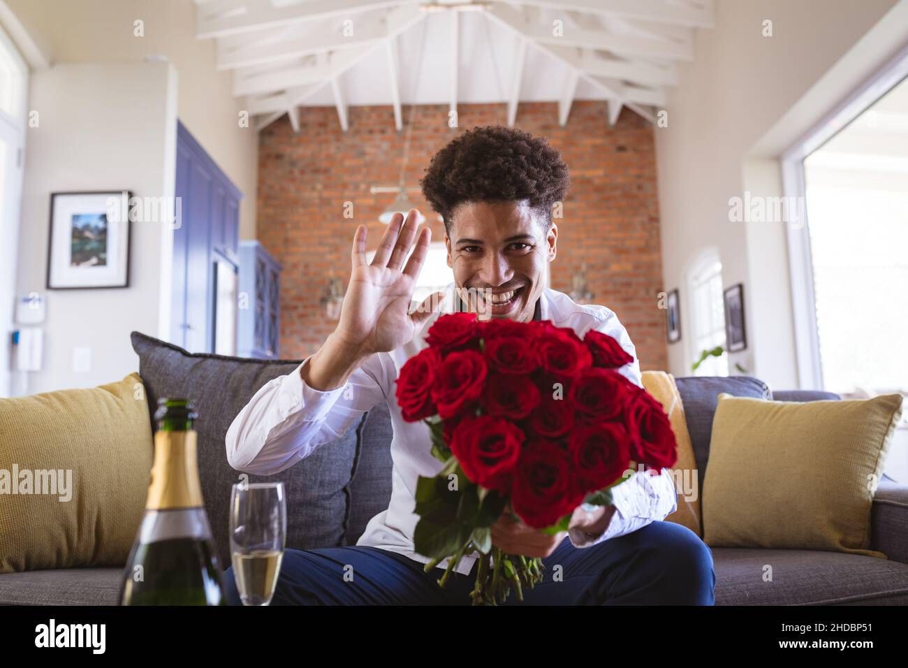 Portrait of happy young man holding rose bouquet waving during virtual date from living room at home Stock Photo