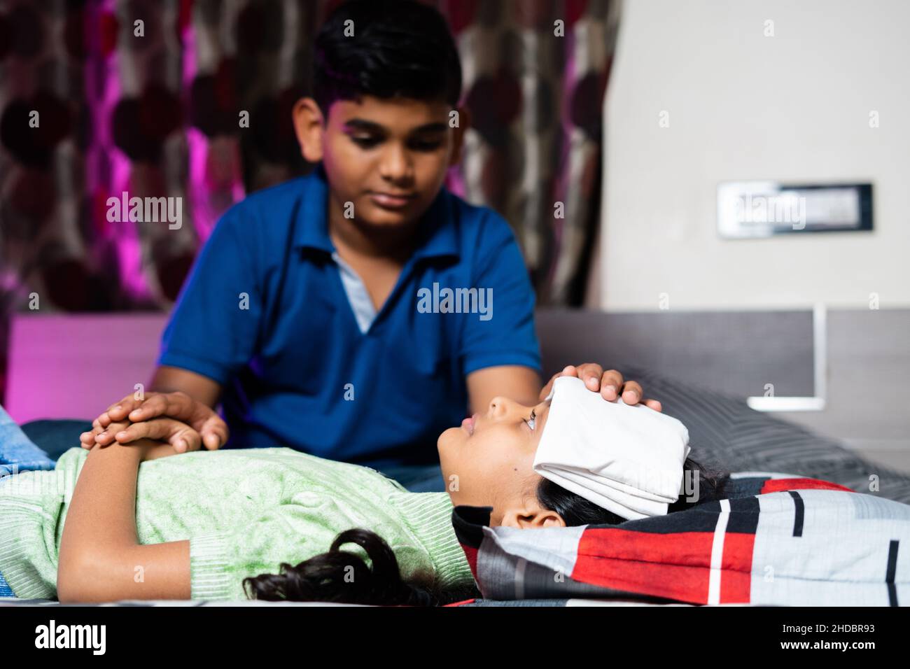 Brother helping by placing cold water cloth on sick sisters forehead due to fever - concept showing childhood sibling caring, relationship and bonding Stock Photo