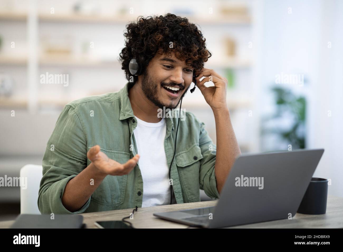 Cheerful indian guy sitting at table, using laptop and headset Stock Photo