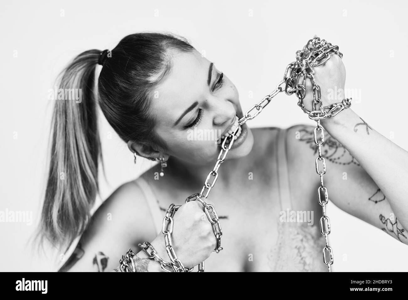 Tattooed feminist woman in bra biting and tearing a iron chain on white background. Girl power concept. Black and white photo Stock Photo