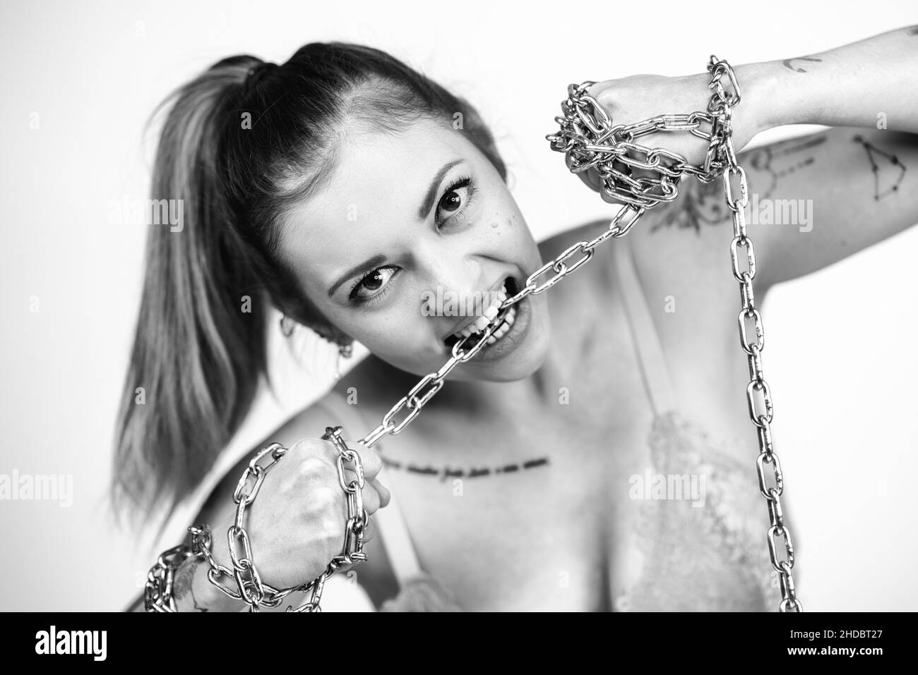 Tattooed woman in bra biting and tearing a iron chain on white background. Girl power concept. Black and white photo. Stock Photo