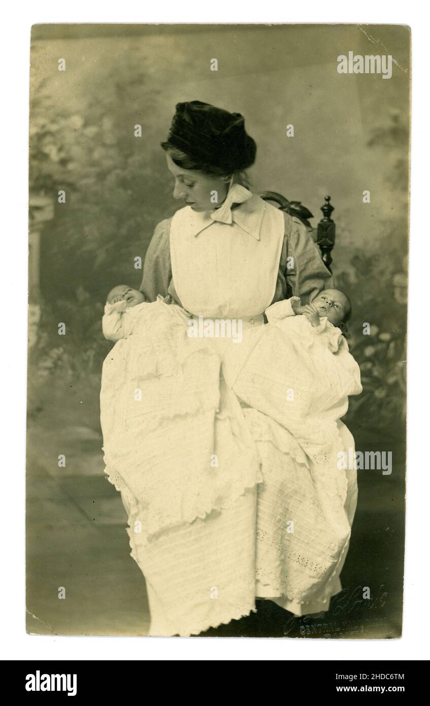 Original early Edwardian era postcard of attractive nanny / nursemaid in starched pinafore uniform holding twins in white christening gowns, Photo by J.L. Clark, Blyth,  Northumberland. Circa 1902 Stock Photo