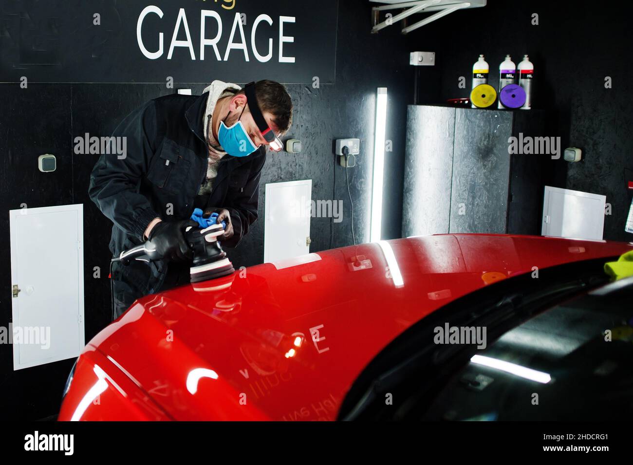 Car detailing concept. Man in face mask with orbital polisher in repair shop polishing orange suv car. Stock Photo