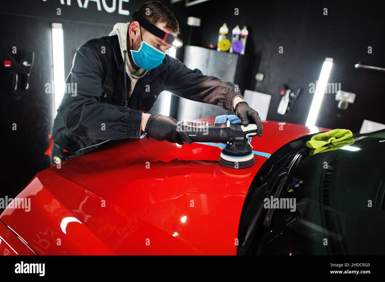 Car detailing concept. Man in face mask with orbital polisher in repair shop polishing orange suv car. Stock Photo