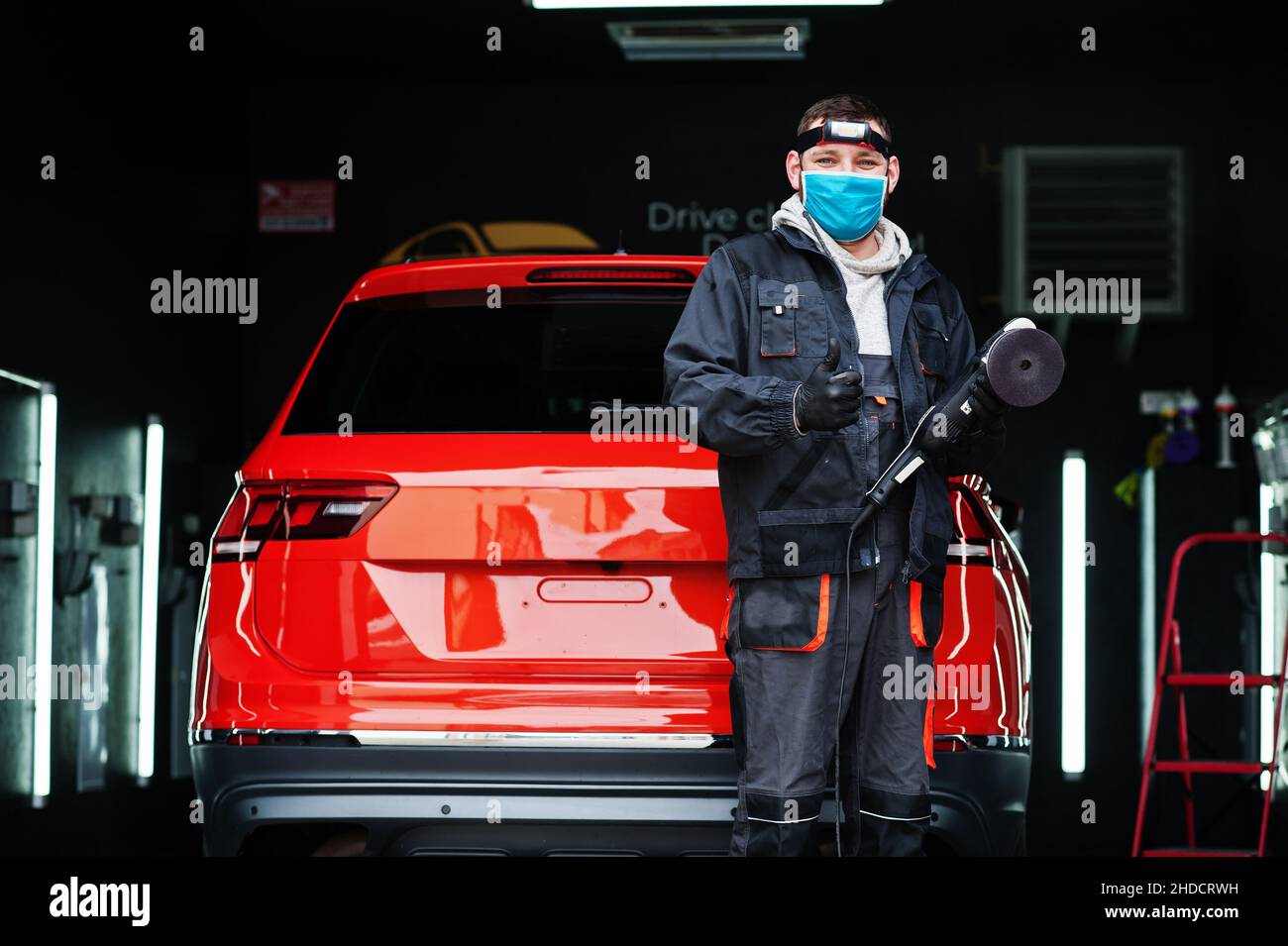 Car detailing concept. Man in face mask with orbital polisher in repair shop polishing orange suv car. Stock Photo