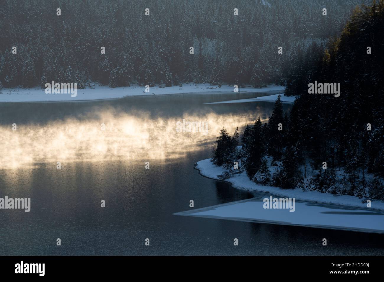 Water vapor in the evening light of sunset on the ice-cold Lake Plansee in winter with a wooded shore Stock Photo