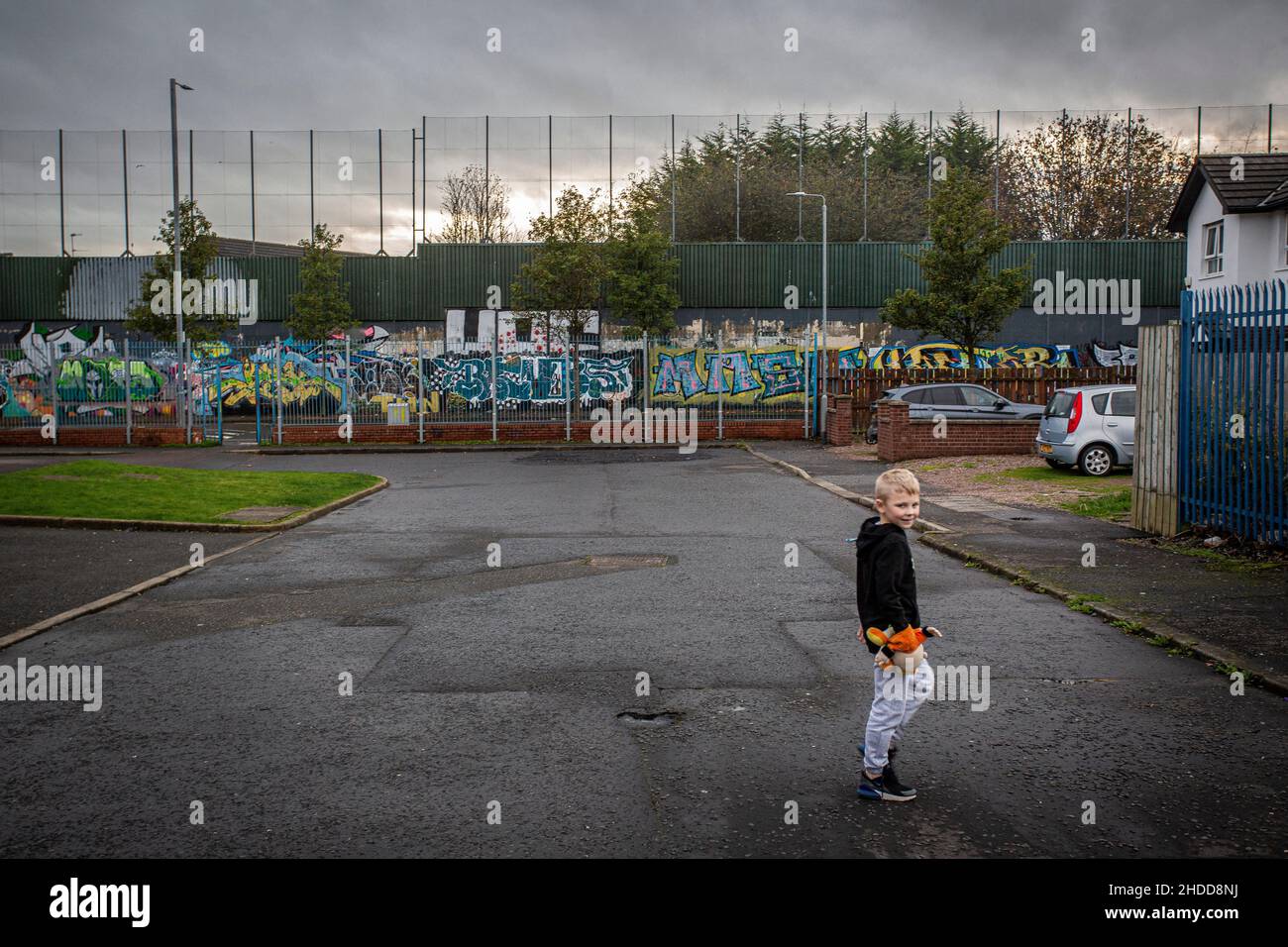 Young boy with colourful murals & graffiti on Peace wall,or Peace Line, running along Cupar Way in Belfast that separate predominantly republican and Stock Photo