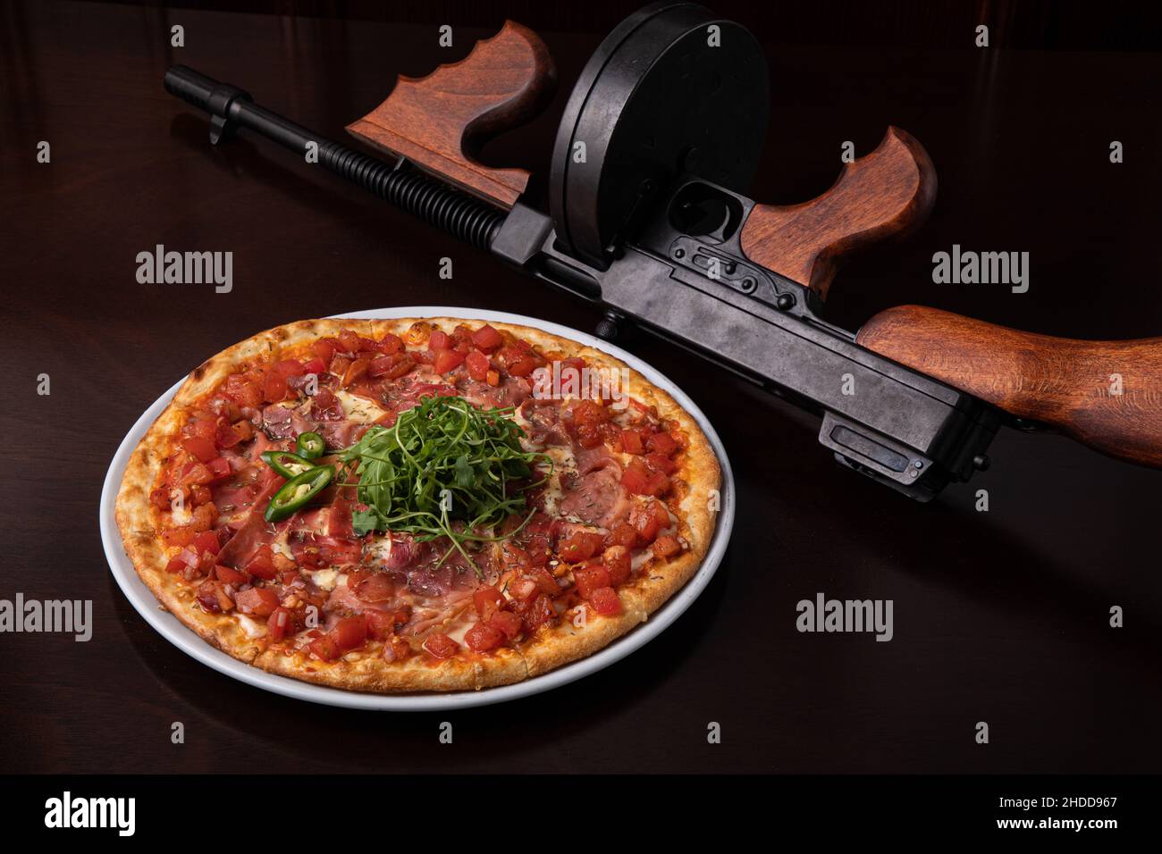 Thompson submachine gun on a dark brown table and a ham pizza with cheese  Stock Photo