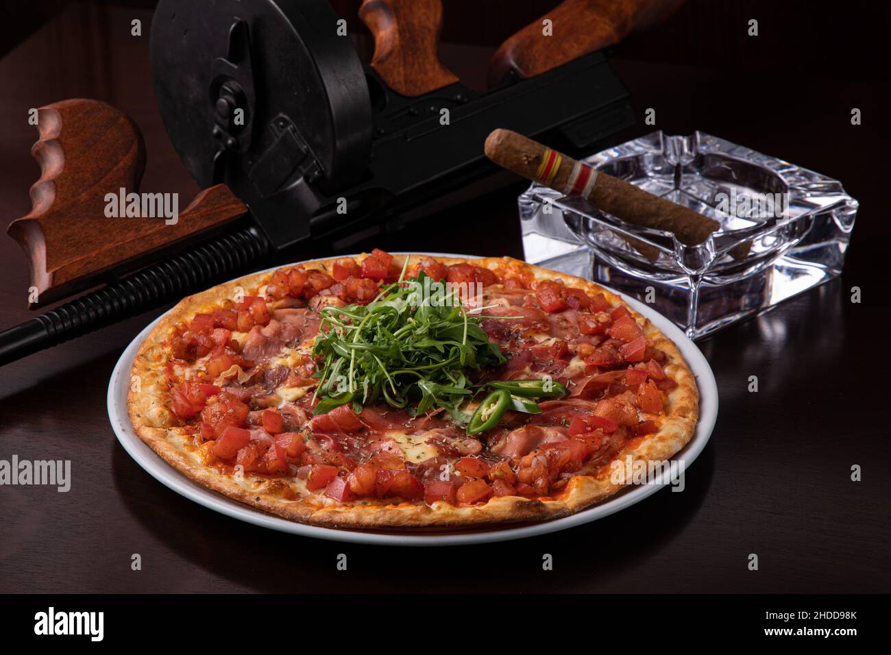 Thompson submachine gun on a dark brown table and ham pizza and a Cuban cigar Stock Photo