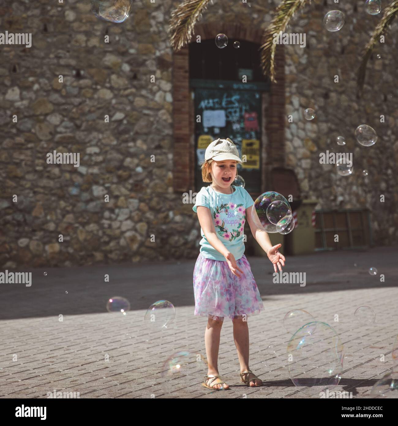 Pretty little girl is playing with big bubbles in street in summer day, Spain Stock Photo