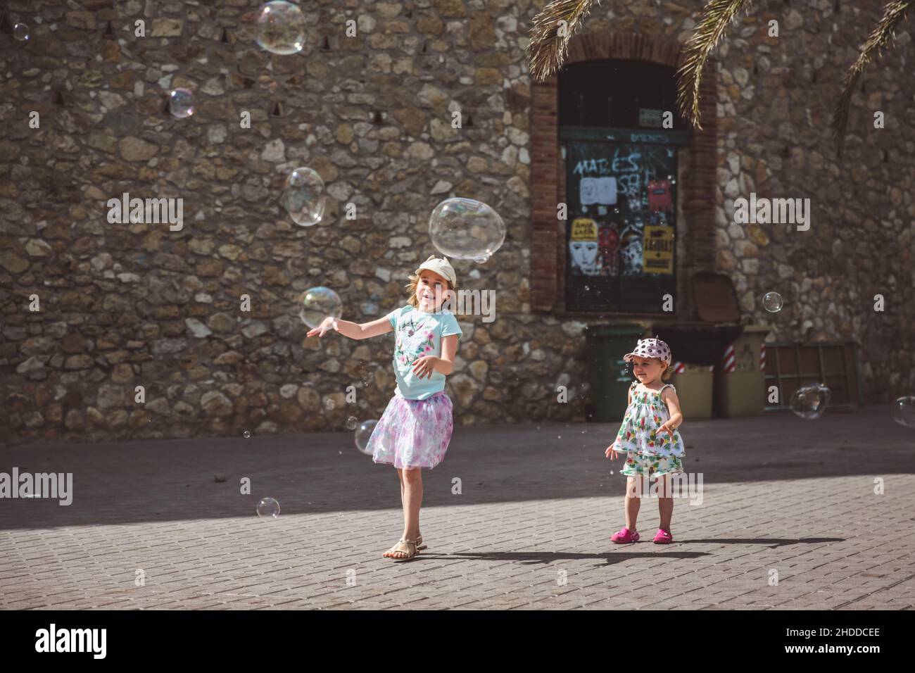 Two pretty little girls are playing with big bubbles in street in summer day, Spain Stock Photo