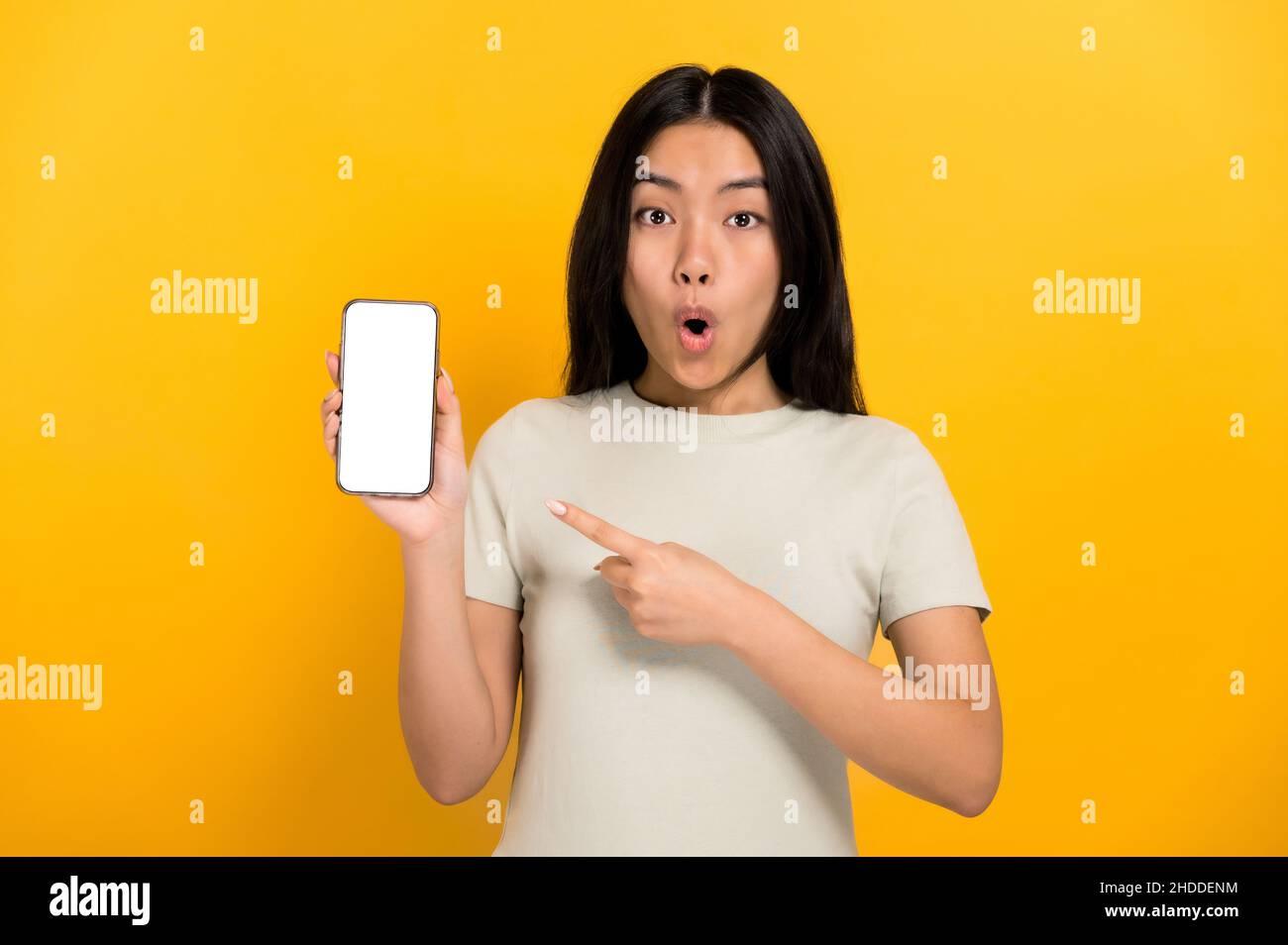 Shocked amazed chinese brunette girl in casual t-shirt showing modern smartphone with blank white screen and points a finger at him, stands on isolated orange background, mockup, copy space Stock Photo
