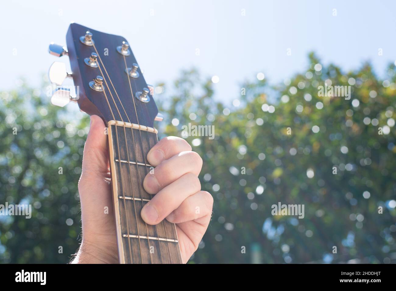 Selective focus of a hand holding an acoustic guitar outdoors, with the sky and greenery in the background Stock Photo