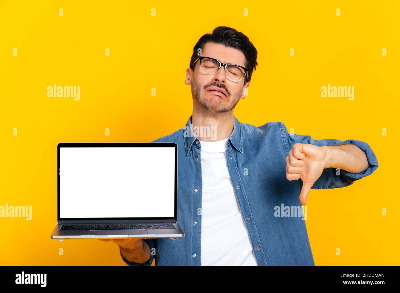 Upset crying caucasian modern guy with glasses, holding open laptop with blank white screen in hand, showing thumbs down, while standing against isolated orange background. Negative emotion Stock Photo