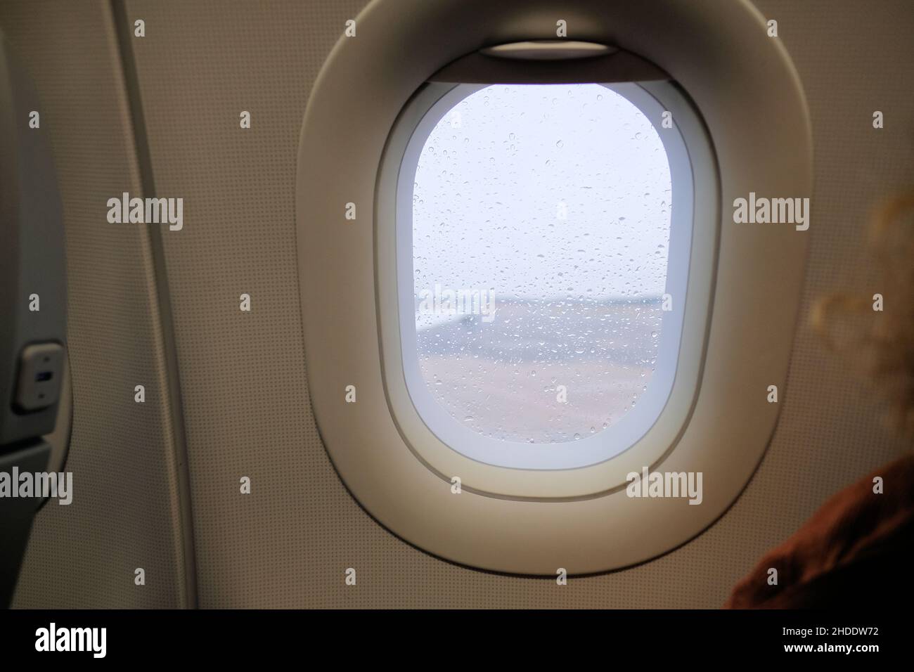Airplane window during a storm with rain drops on window pane; selective focus on raindrops with blurry tarmac, wing, and gray exterior. Stock Photo