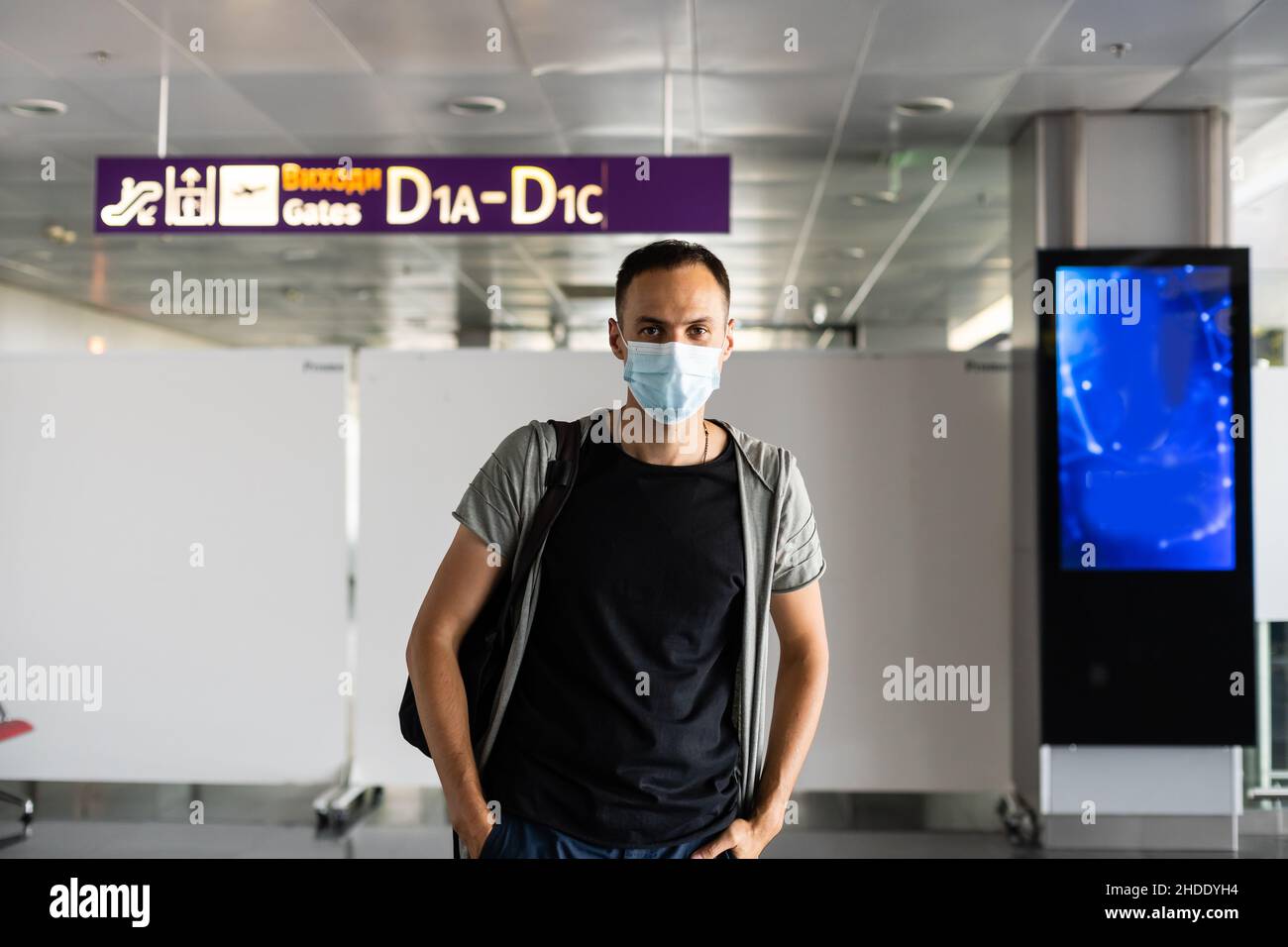 Young European man in gray coat, protective disposable medical mask in airport. Afraid of dangerous N-CoV 2019 influenza coronavirus. backpack Stock Photo