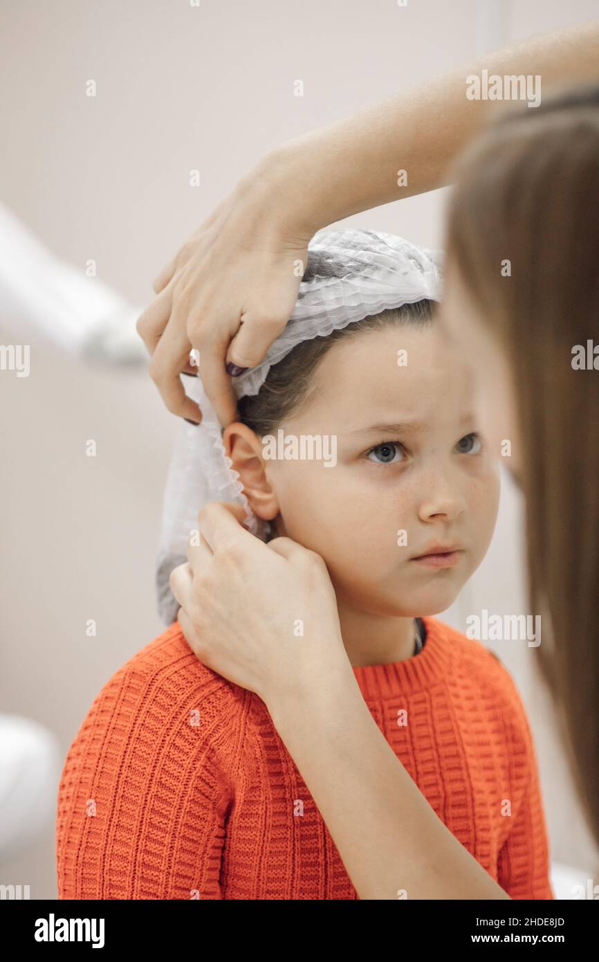 close-up of beautician hands putting white disposable medical cap on head of six-year-old girl in cosmetology office, preparing for ear piercing Stock Photo