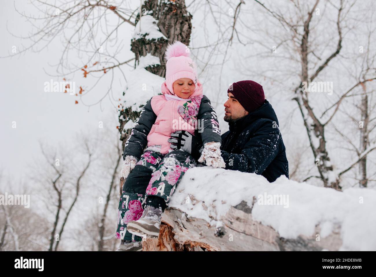 Distant photo of female kid sitting on tree log and smiling wearing pink winter clothes with father holding daughter in forest. Astonishing background Stock Photo