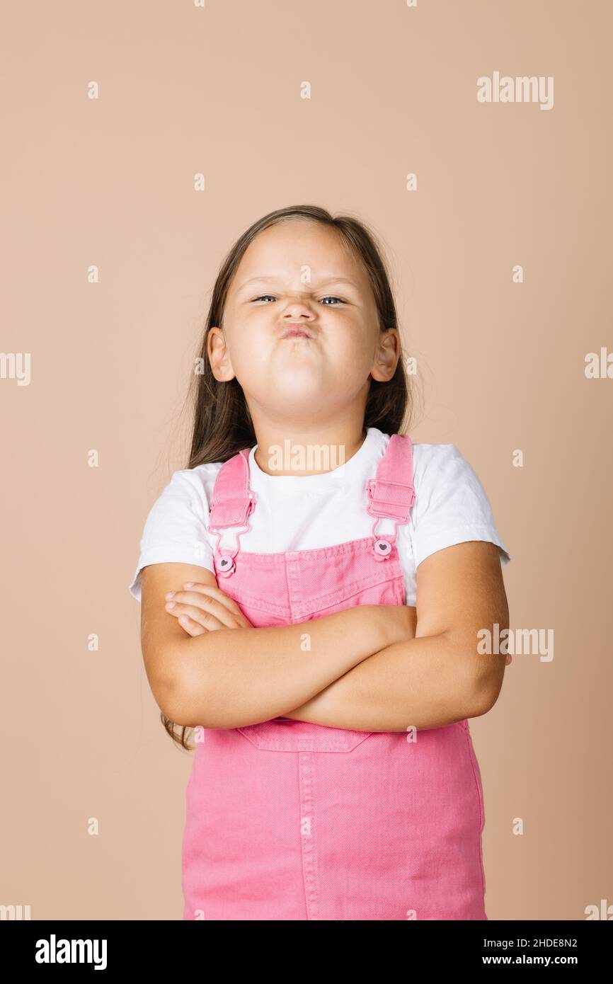 Portrait little girl with folded arms with dissatisfied grimacing face looking at camera wearing bright pink jumpsuit and white t-shirt on beige Stock Photo