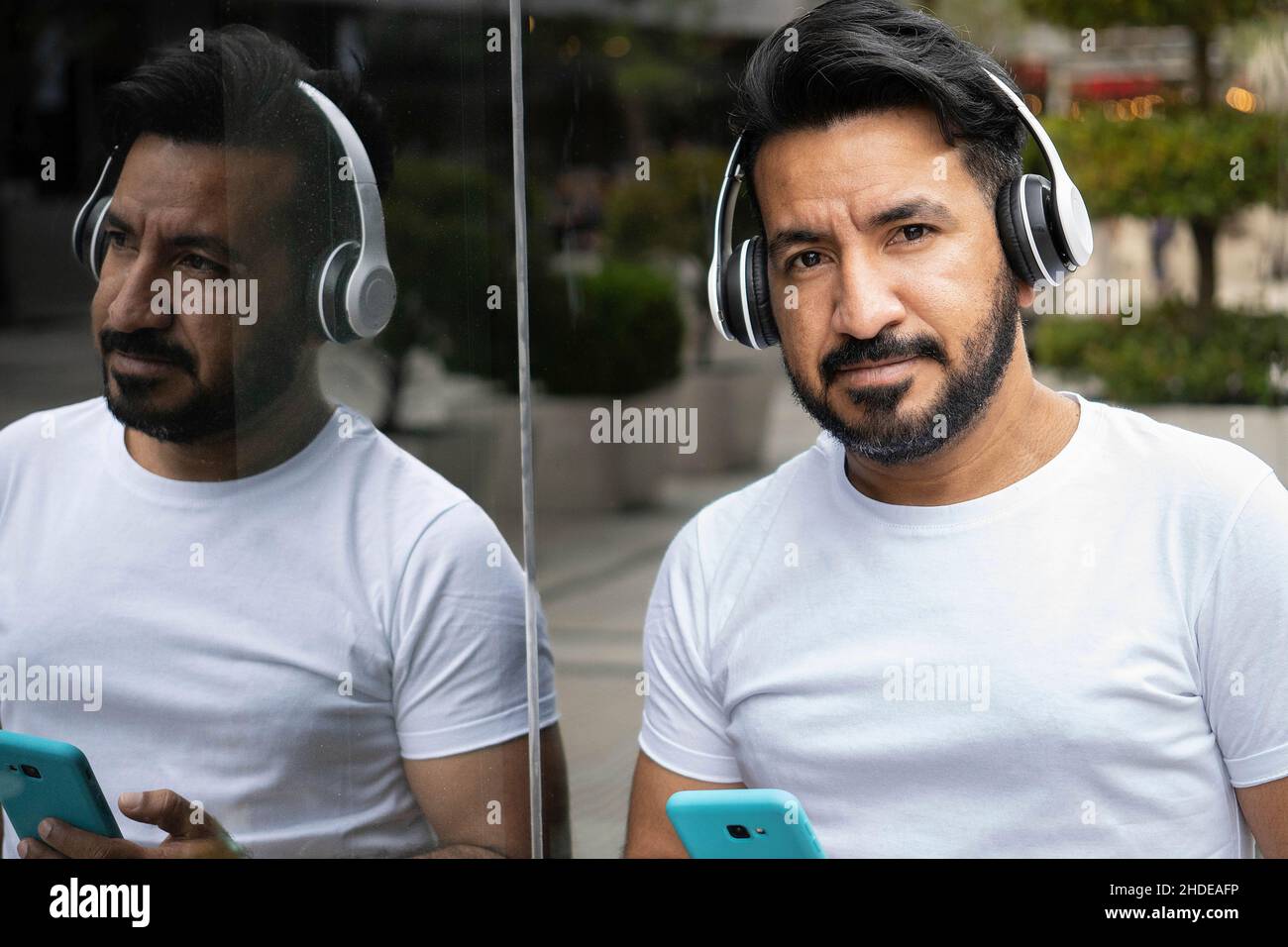 latin man with beard reflected in the glass of a building with cell phone and headphones Stock Photo