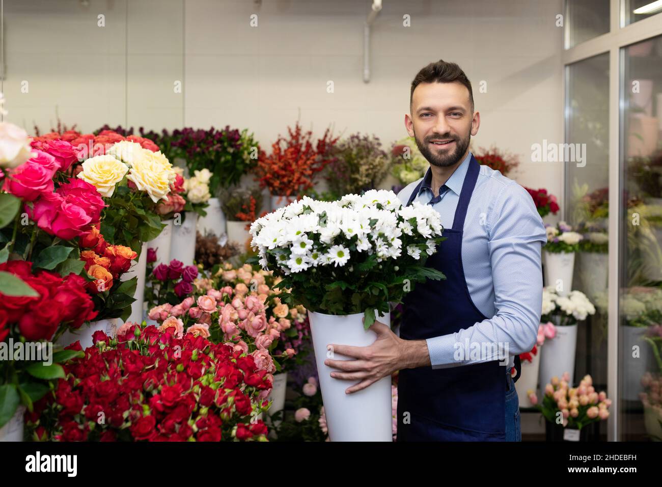 a man florist in the refrigerator among fresh flowers holding a vase with live plants in his hands looks at the camera with a smile Stock Photo