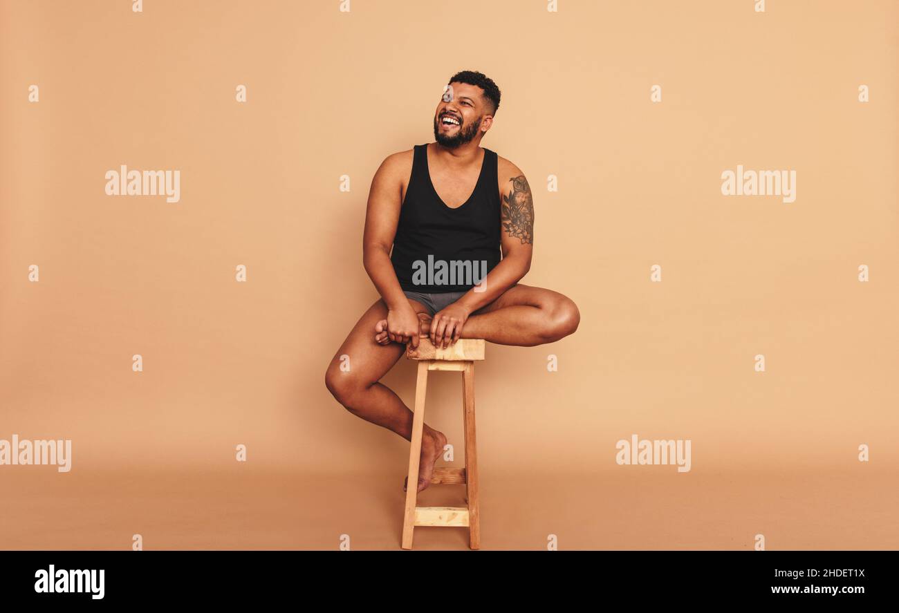 Tattooed man smiling cheerfully while sitting on a wooden chair in a studio. Body positive young man looking away while sitting alone. Self-confident Stock Photo