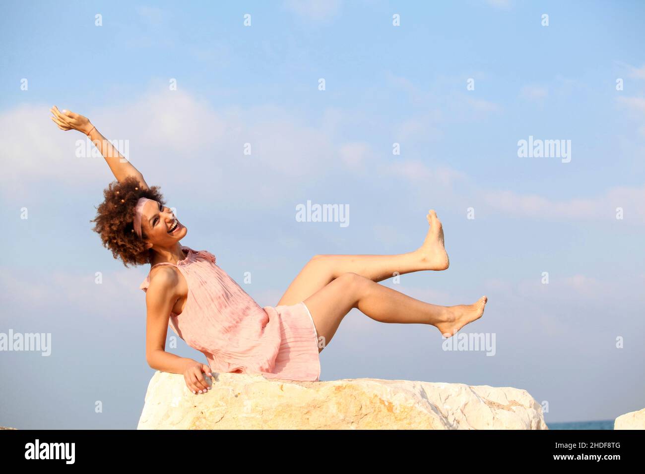 A happy, smiling 28 year old new age style woman sits on a rock on the beach Stock Photo