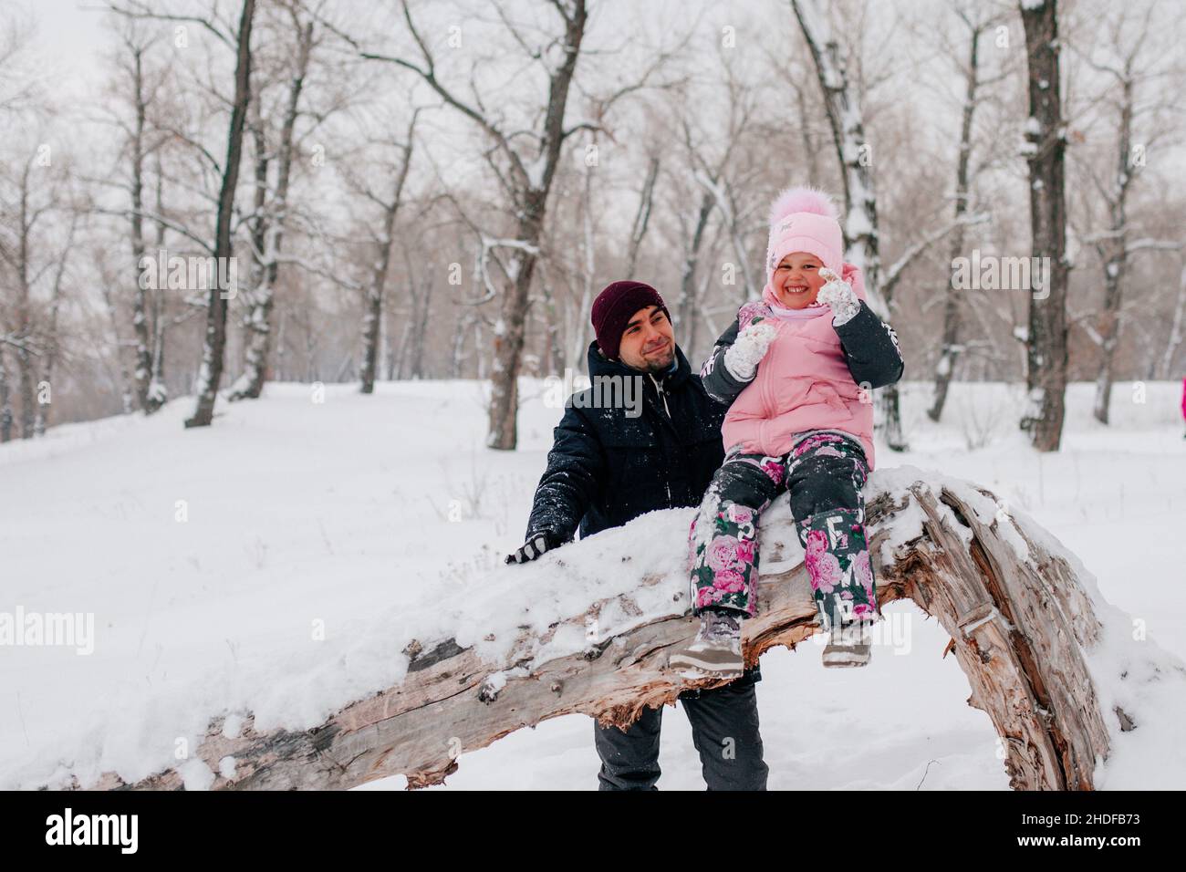 Distant photo of female kid sitting on tree log super excited wearing pink winter clothes with father standing behind daughter in forest. Astonishing Stock Photo