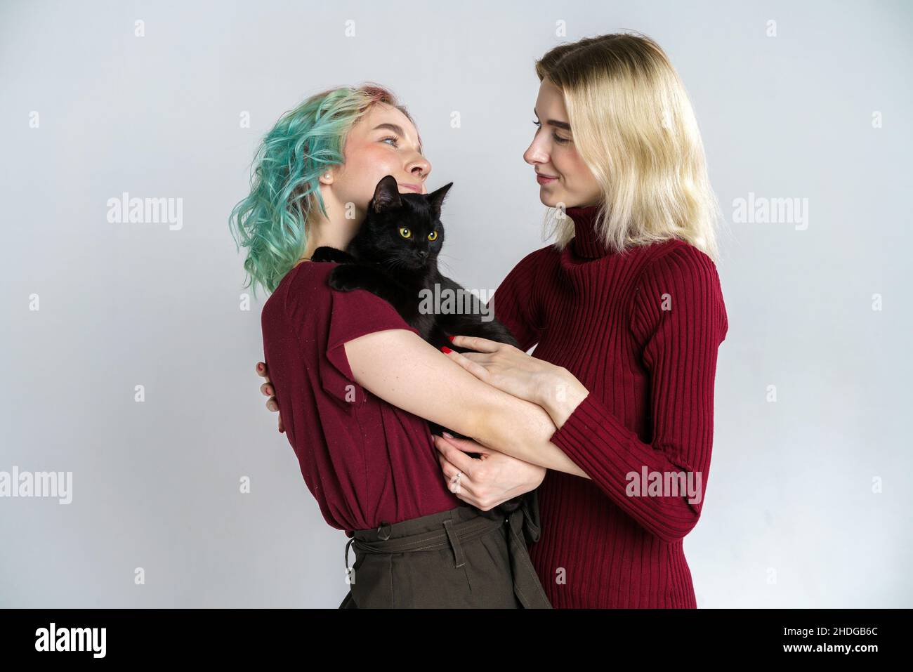 Portrait happy girls in casual clothes on light background, holding black cat in their hands and hugging him, smiling and kissing. Cheerful friends hug the cat and rejoice. Stock Photo