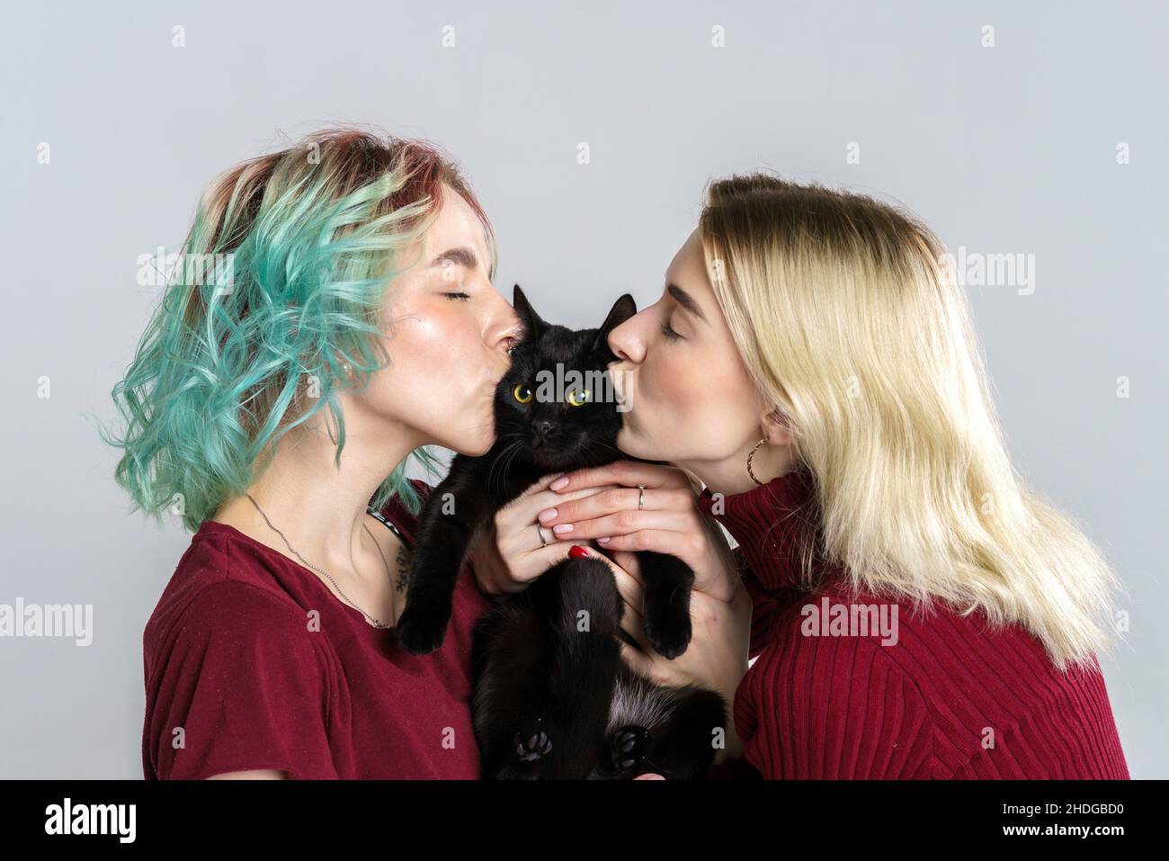Portrait happy girls in casual clothes on light background, holding black cat in their hands and hugging him, smiling and kissing. Cheerful friends hug the cat and rejoice. Stock Photo