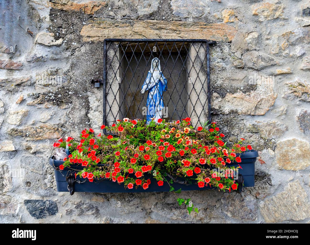 statue of the Virgin Mary in the niche of a stone wall behind an iron grid and a jardiniere with red blossoms, Austria Stock Photo