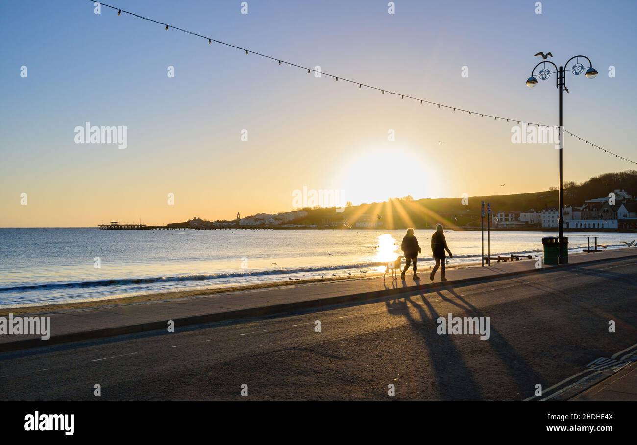 Dog walkers walking along pavement at Swanage bay early morning during a sunrise with blue sky and sun star over Swanage hill near Peveril point. Stock Photo