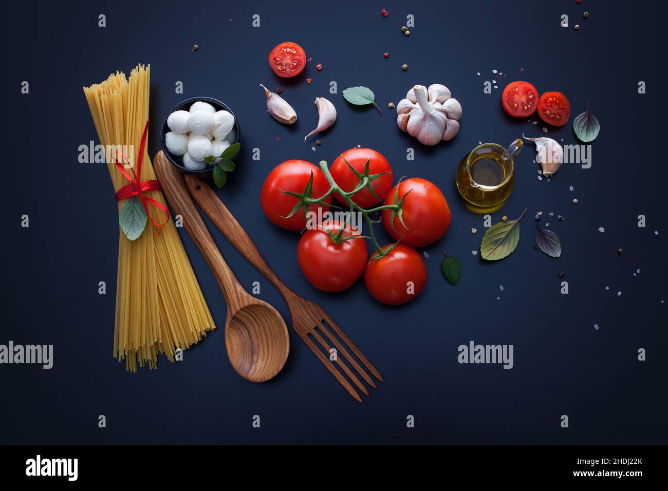 A vibrant spread of fresh garlic, basil, cherry tomatoes, and mozzarella pearls surrounding a pile of spaghetti.