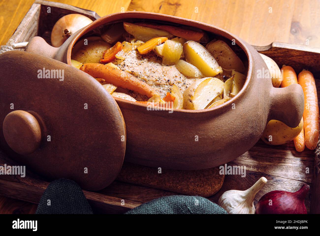 Side view of terracotta clay cooking pot with slow cooked pork roast and vegetables inside on wood tray and wooden table, surrounded by raw organic. Stock Photo