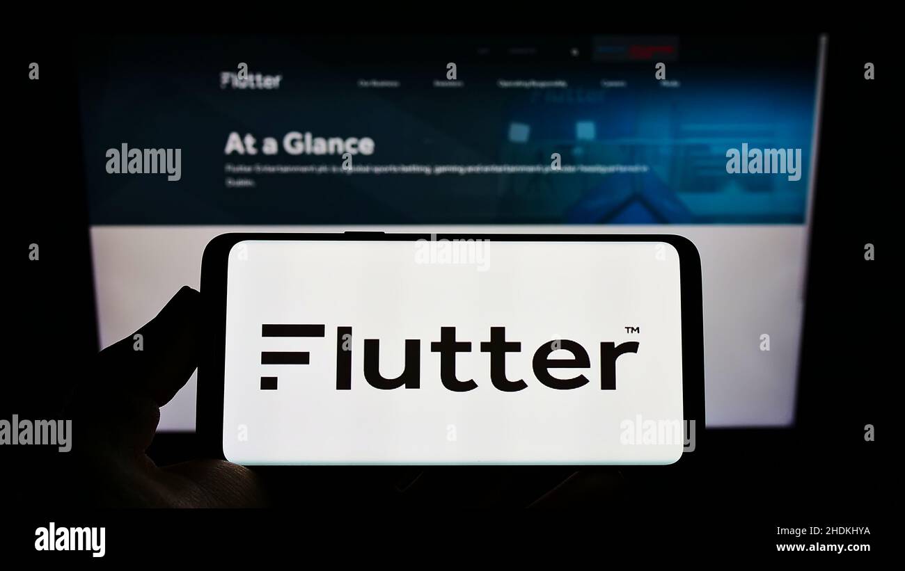 person holding mobile phone with logo of irish bookmaking company flutter entertainment plc on screen in front of web page focus on phone display 2HDKHYA