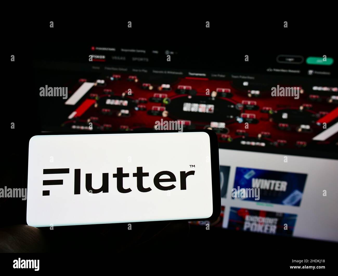 Person holding cellphone with logo of bookmaking company Flutter Entertainment plc on screen in front of Pokerstars website. Focus on phone display. Stock Photo