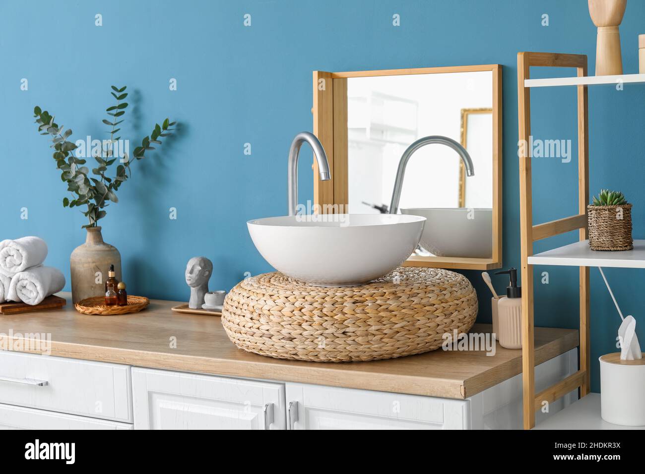 Rattan stand with sink, mirror and bath supplies near blue wall Stock