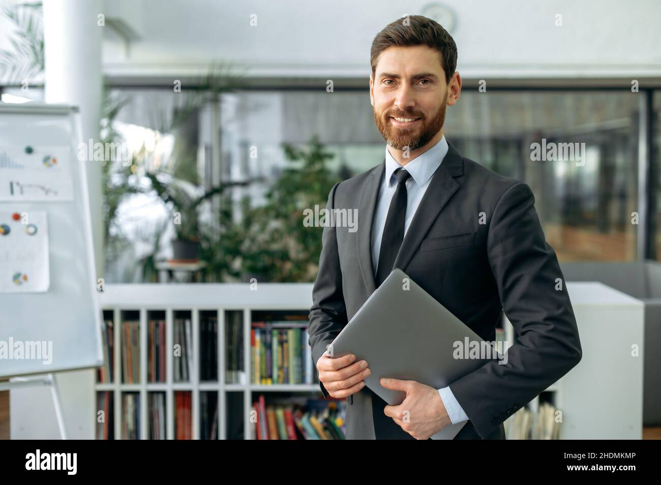 Portrait confident intelligent proud caucasian businessman, top manager, lawyer or IT specialist, in shirt, stands in modern office, holding a laptop, looking at the camera with a friendly smile Stock Photo