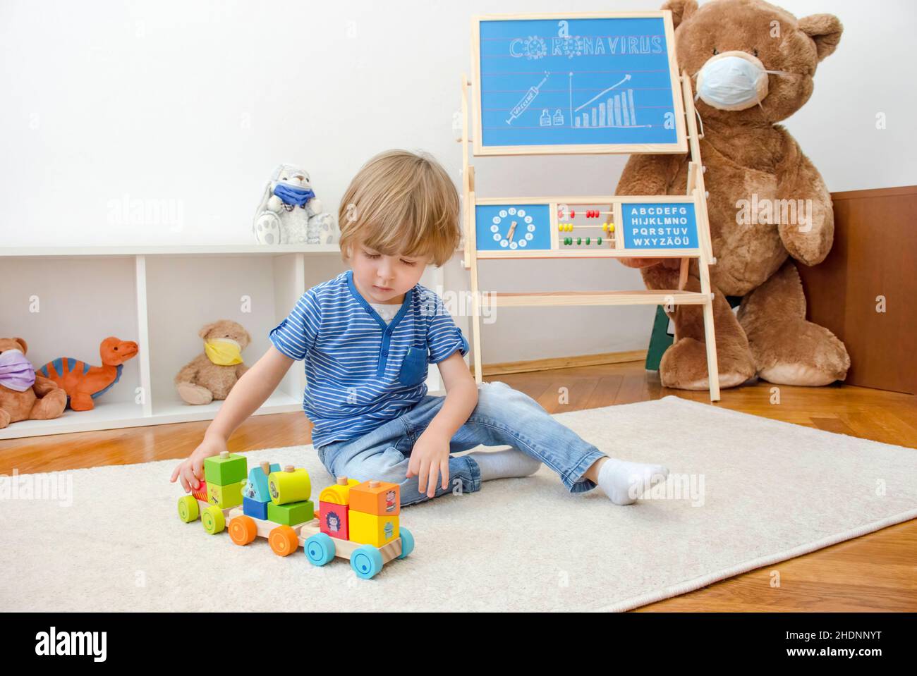 Cute child boy playing with wooden toys at home on the floor and the growing graph of COVID-2019 cases drawn on a chalkboard, during coronavirus pande Stock Photo