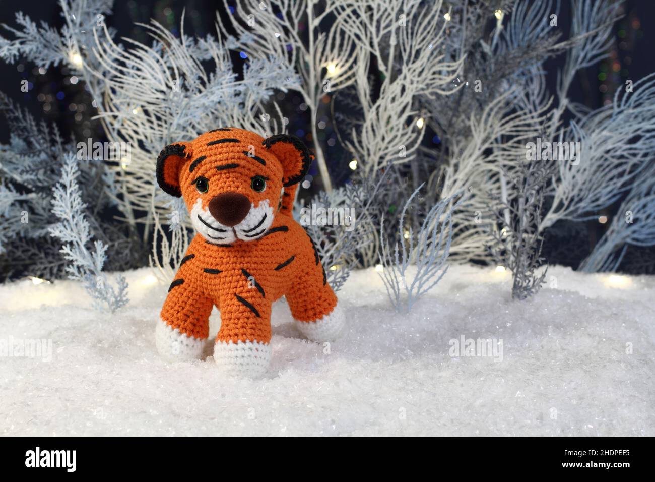 A knitted toy tiger stands on artificial snow and artificial blue and white trees with festive lights stand behind them against a dark blue background Stock Photo