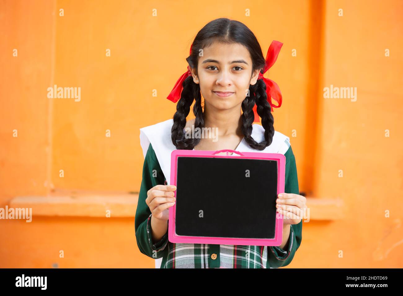 Young beautiful happy indian school girl holding slate against orange background, Smiling braided hair female teenager kid with black board. education Stock Photo