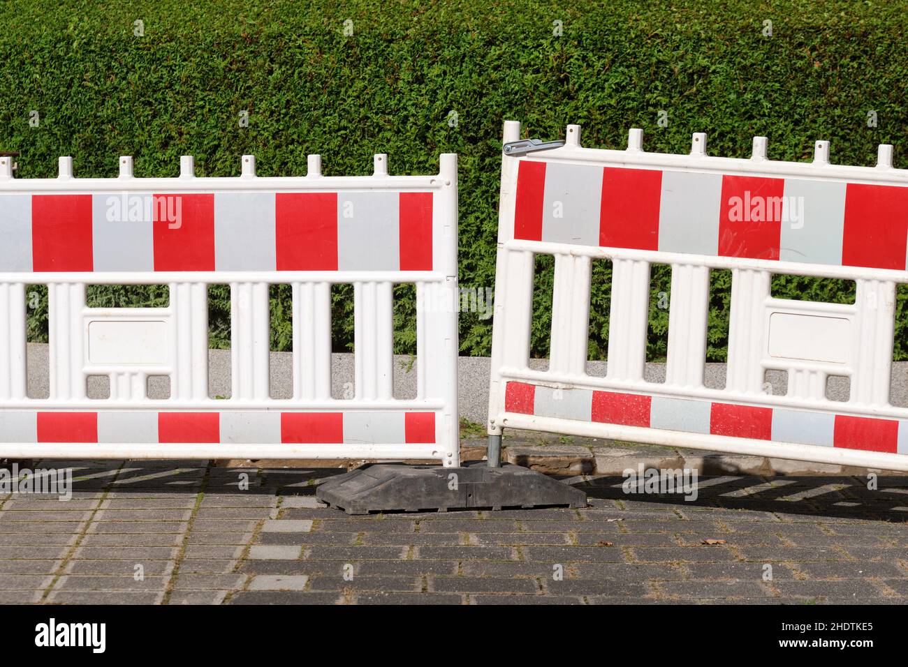 Red and white street barriers standing in front of a construction site on the pavement in front of a green hedge. Seen in German in October. Stock Photo