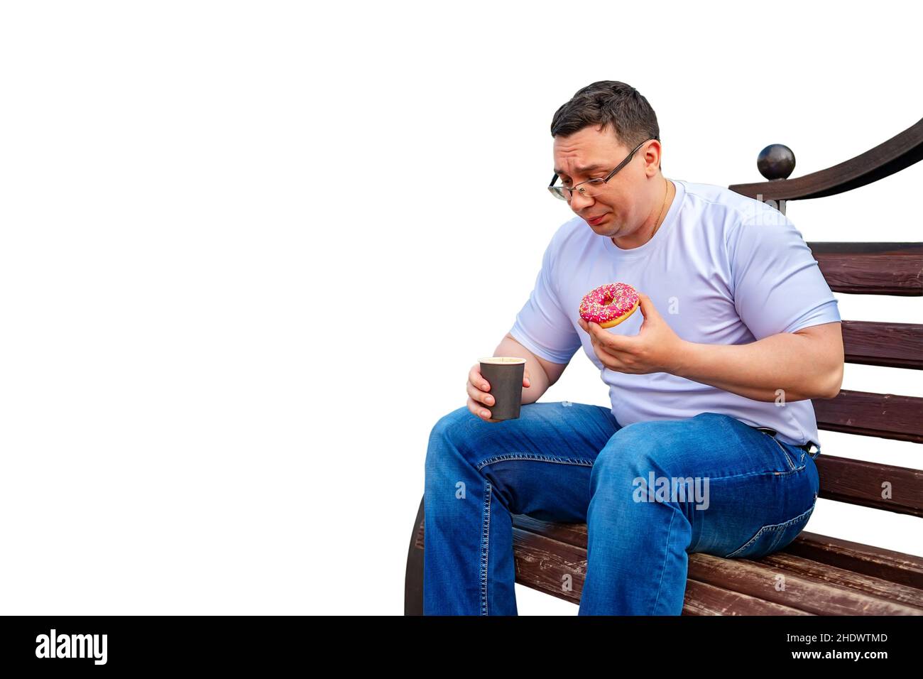 a sad, puzzled, sad man with glasses is sitting on a bench, having lunch, eating a sweet donut, a bun and holding a cup of coffee, tea Stock Photo
