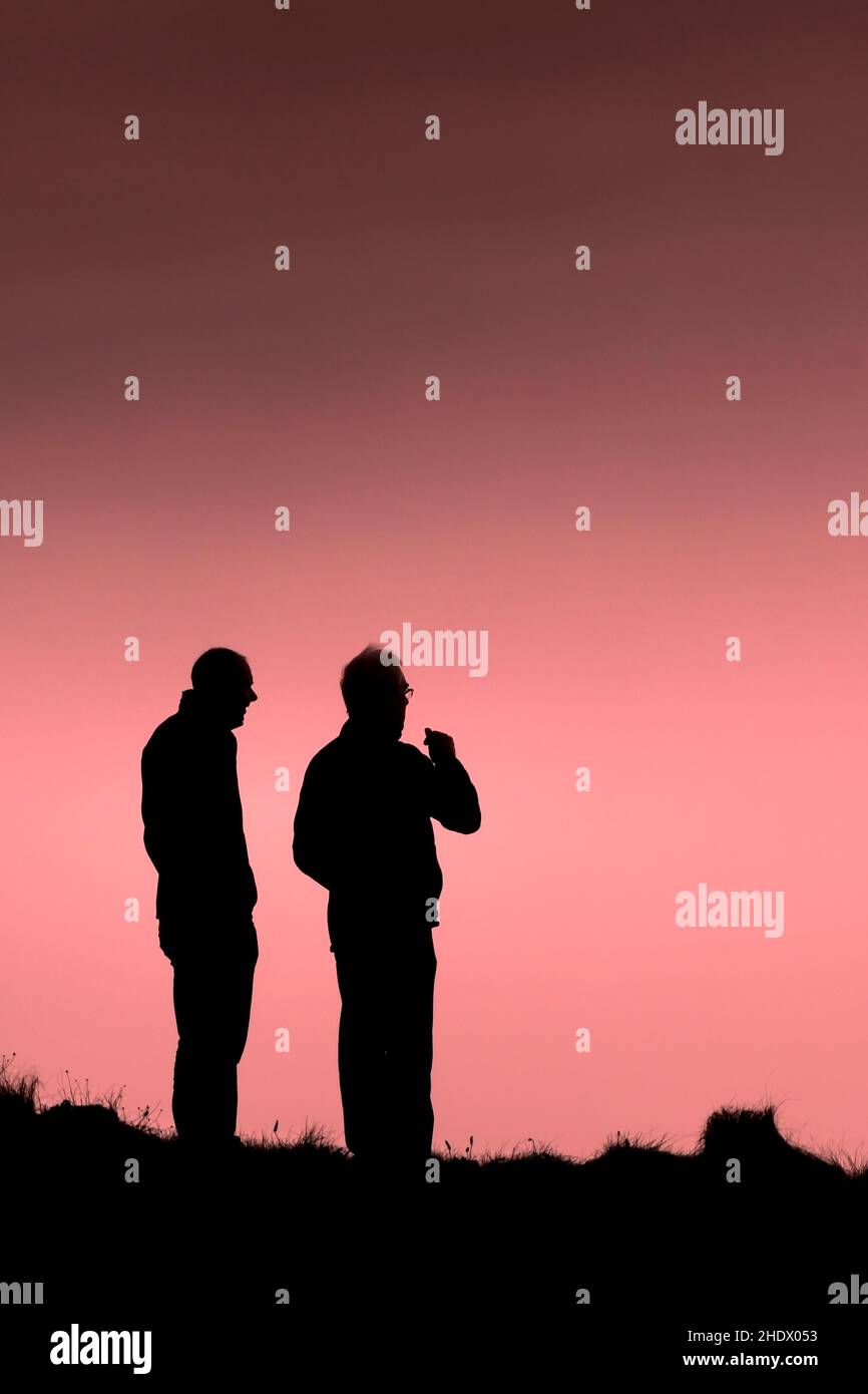 Two people standing on the coast path silhouetted against a colourful sky at the end of the day in Newquay in Cornwall. Stock Photo