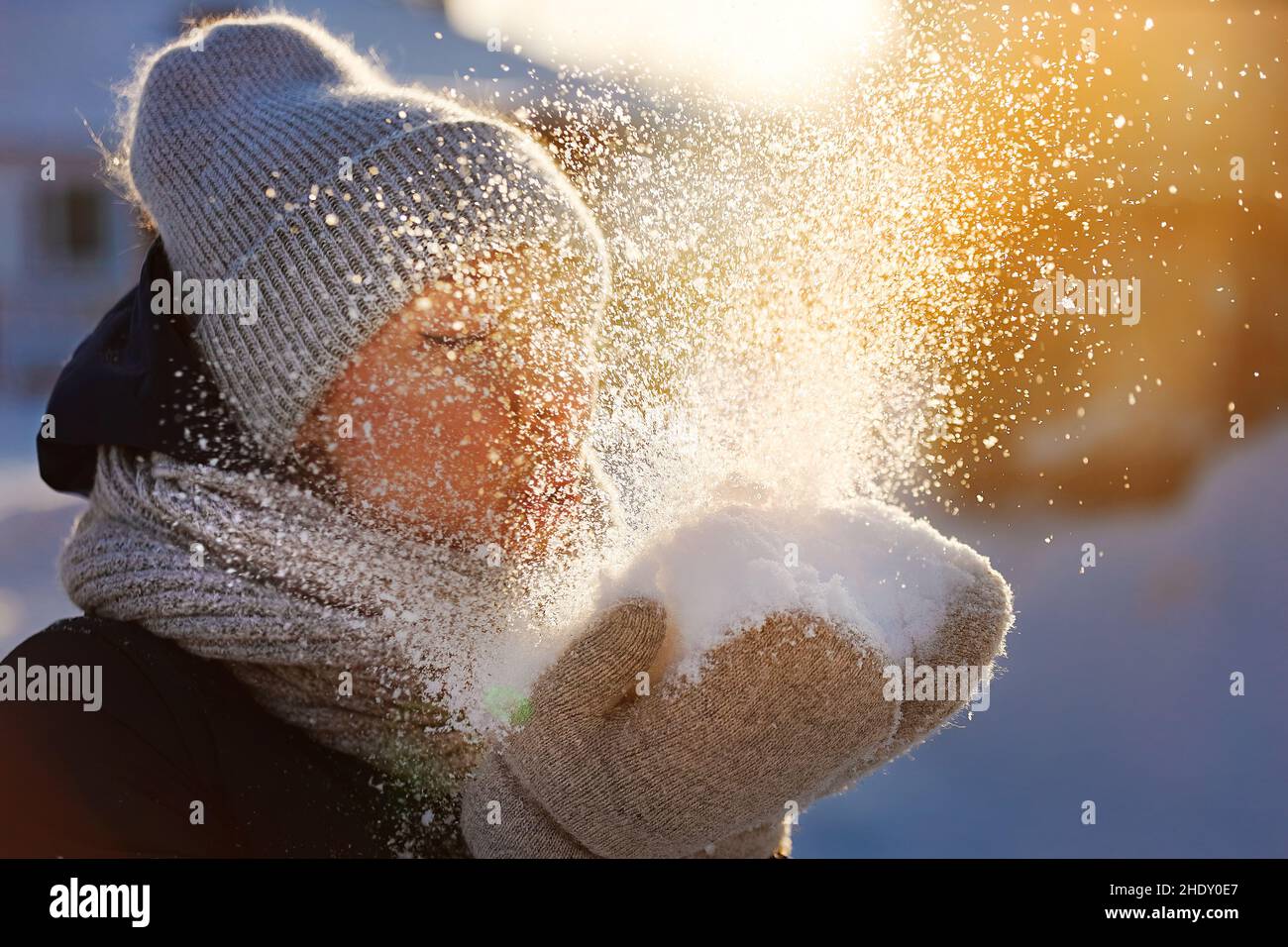 Girl in winter clothes blows on snow from her palms and snowflakes fly. Winter mood. Happy woman of 35-40 years old has fun on clear frosty day. Sun glare. Stock Photo