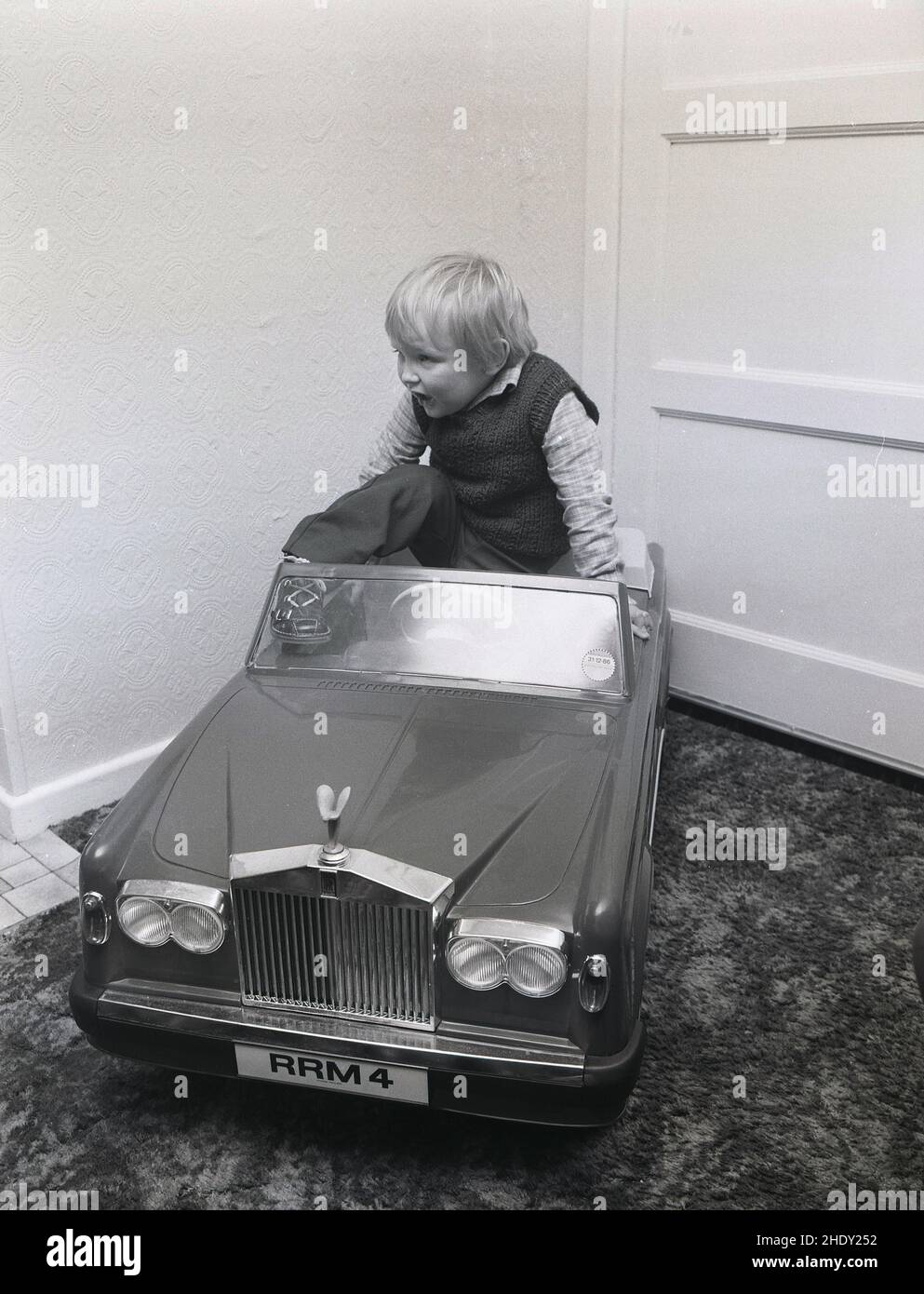 1980s, historical, inside a room, a little boy climbing into the seat of his ride-on toy car, a Rolls Royce Corniche with personalised numberplate RRM 4, England, UK. This classic child's pedal car was made by Triang, a leading British toy maker of the day. It had a moulded plastic body on a steel frame and an electric motor. As a young boy, Prince WIlliam had one given to him by Harrod's, the famous store in London's Knightsbridge which originally sold them. Stock Photo