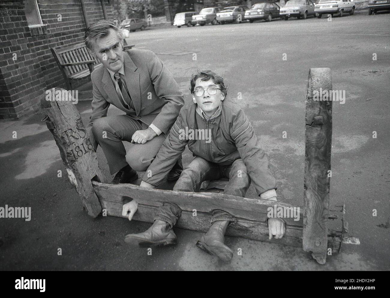 1986, historical, outside a school building, a male teacher and teenage boy demonstrate an ancient wooden 'foot and hand lock', known as the stocks, an instrument of corporal punishment, used to restrain and humiliate offenders. A key part of the stocks was the element of public punishment. It's last recored use in Britain was in 1872. The stocks differed from the pillory, which locked the head of the offender forcing them to stand and where custom dictated that their head and beard were shaved. Stock Photo