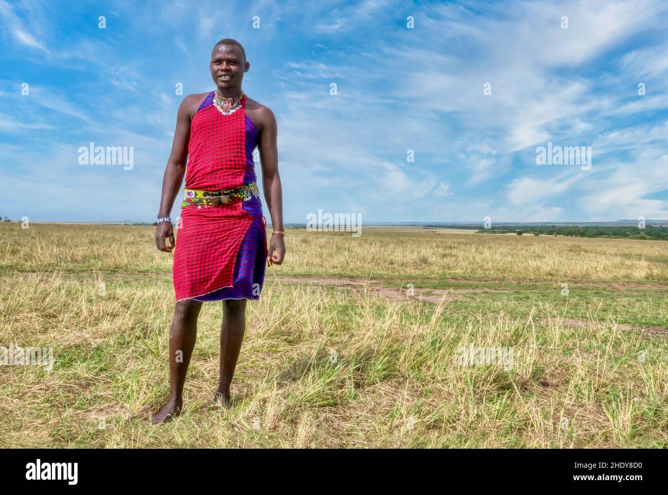 Masai Mara, Kenya - Sept. 29, 2013. A male member of the Maasai tribe wears traditional clothing and jewelry, with a wide shot of the landscape. Stock Photo